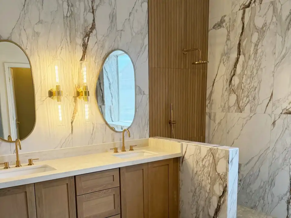 Bathroom remodeling Peoria AZ: marble-look porcelain slab walls, wood double vanity, oval mirrors and brass sconces