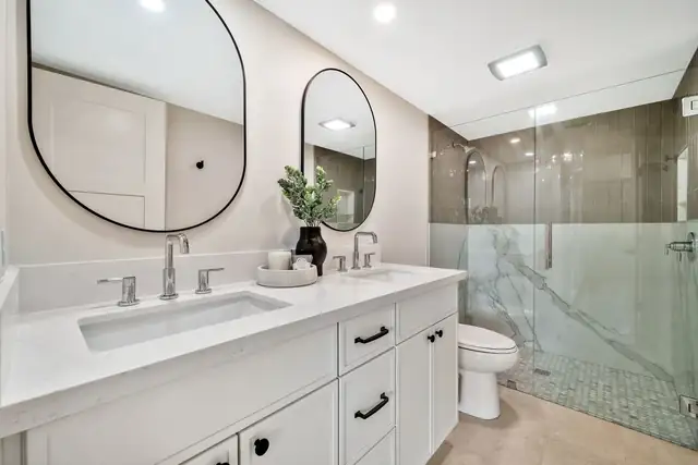 Bathroom remodel with white shaker vanity, oval black mirrors and a marble-look shower with glass door