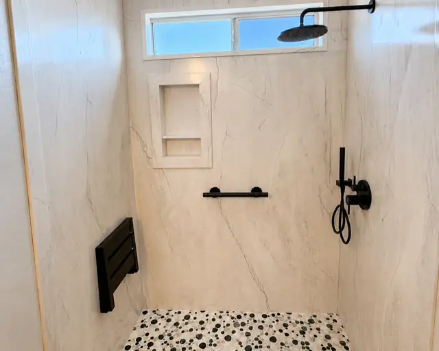 Accessible walk-in shower with a fold-down seat, black grab bar, recessed niche and pebble tile floor